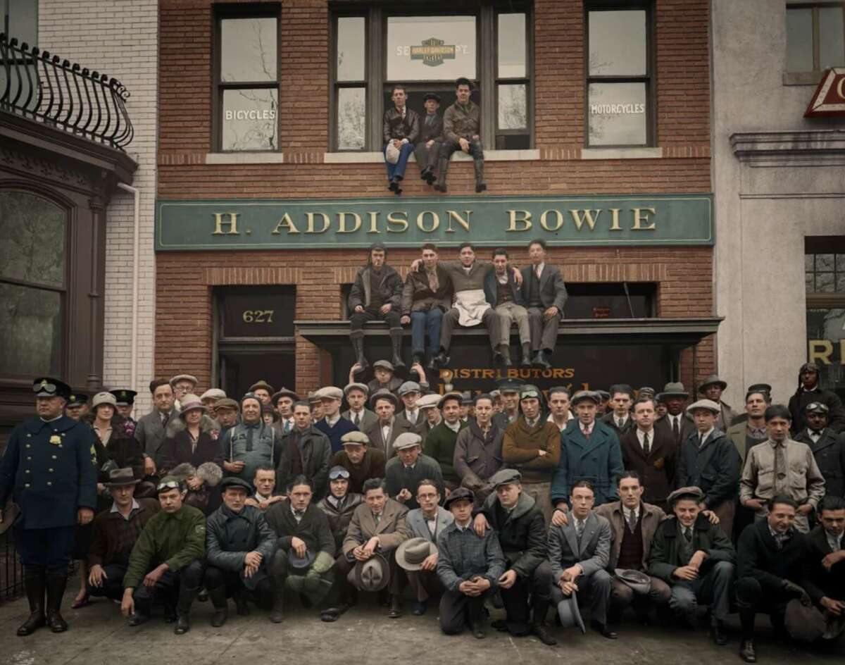 A large group of men in early 20th-century clothing pose in front of a brick building labeled "H. Addison Bowie." Some sit on the steps and balcony, while a policeman stands to the left.