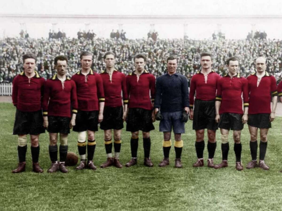 A vintage photo of a soccer team in red jerseys, black shorts, and long socks stands in a row on a grassy field, with a packed stadium of spectators in the background.