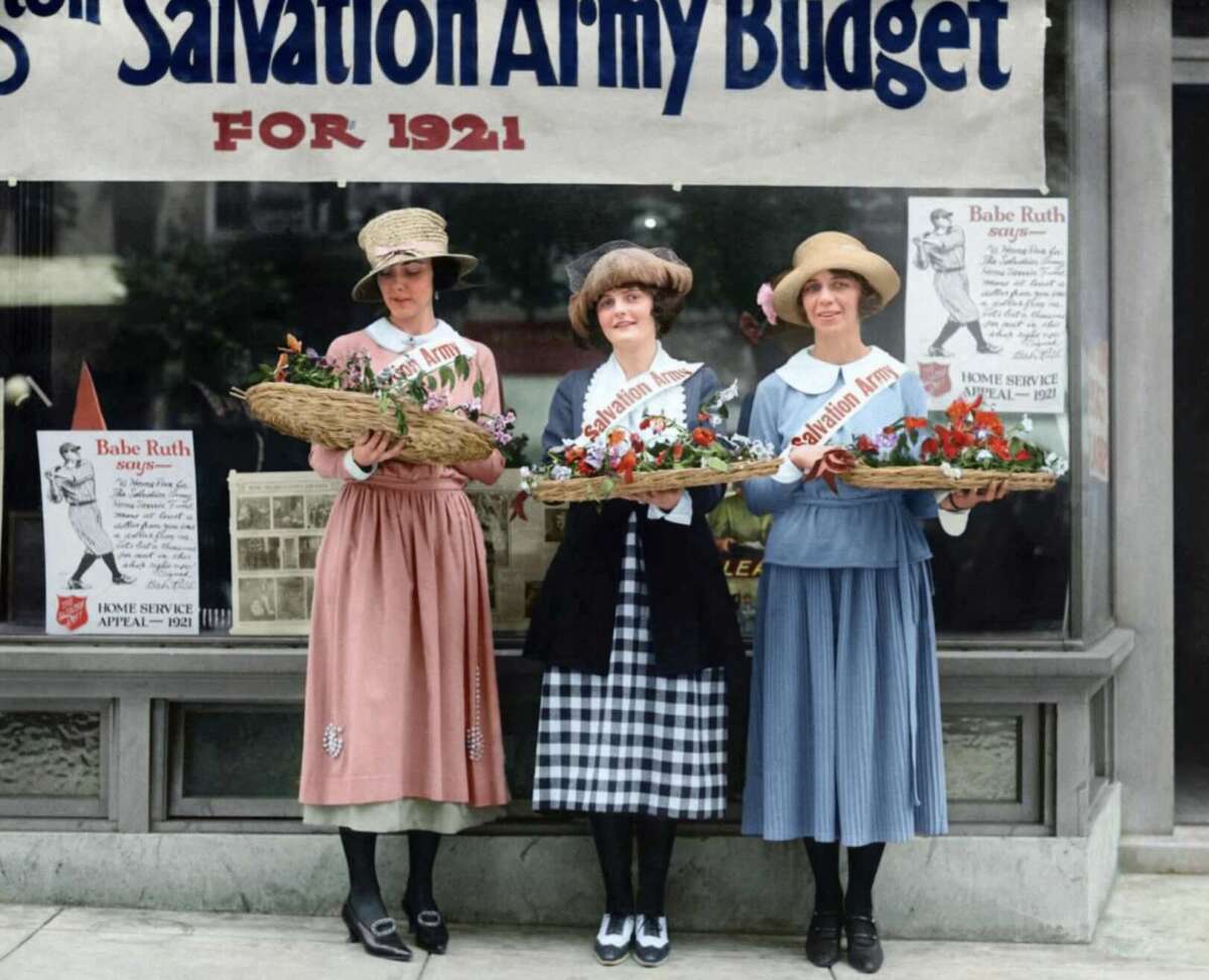 Three women in early 1920s clothing stand outside a Salvation Army window display, each holding a basket of flowers. The sign behind them reads "Salvation Army Budget for 1921." Posters of Babe Ruth are also visible.