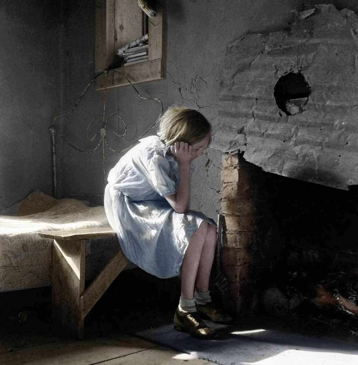 A young girl in a light blue dress sits on the edge of a bed, covering her face with her hands, next to a damaged wall and fireplace in a dim, worn room with minimal furnishings.