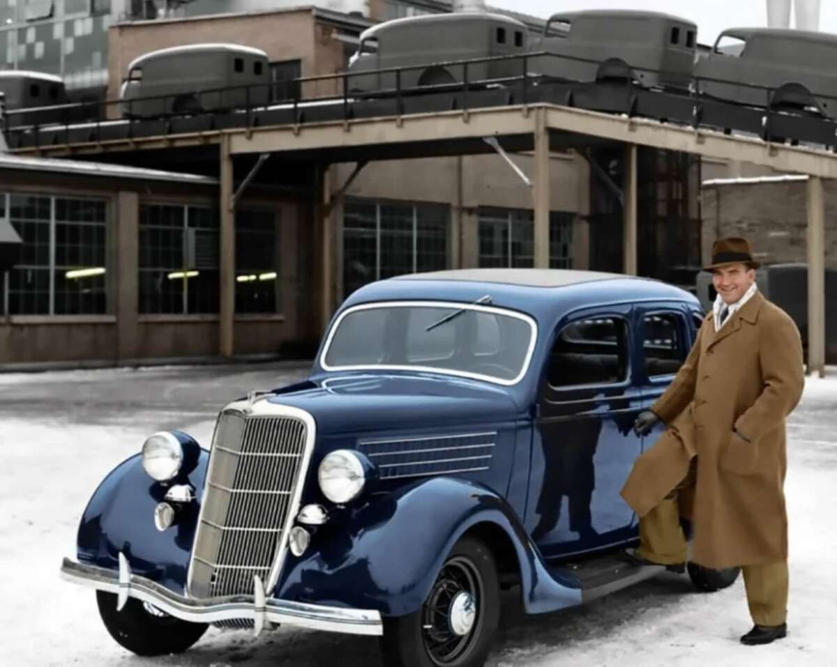 A man in a brown coat and hat stands beside a shiny blue vintage car on a snowy street, with an industrial building and several car bodies stacked on a platform in the background.