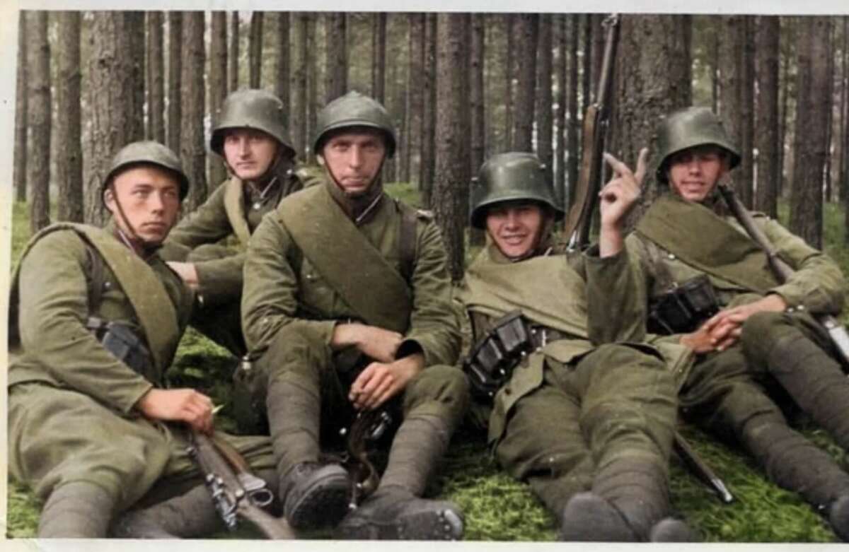 Five soldiers in green military uniforms and helmets sit on grass in a forest, posing for the photo. Some hold rifles and ammunition pouches, and tall trees are visible in the background.
