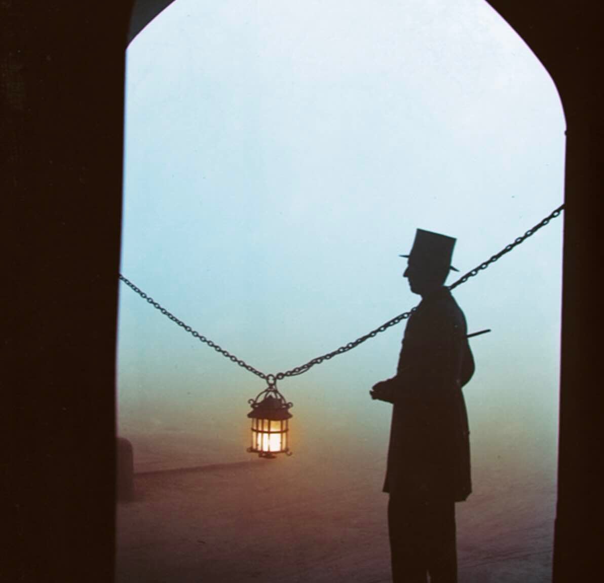 A silhouette of a person in a top hat stands under an archway near a hanging lantern, with fog or mist creating a hazy background.