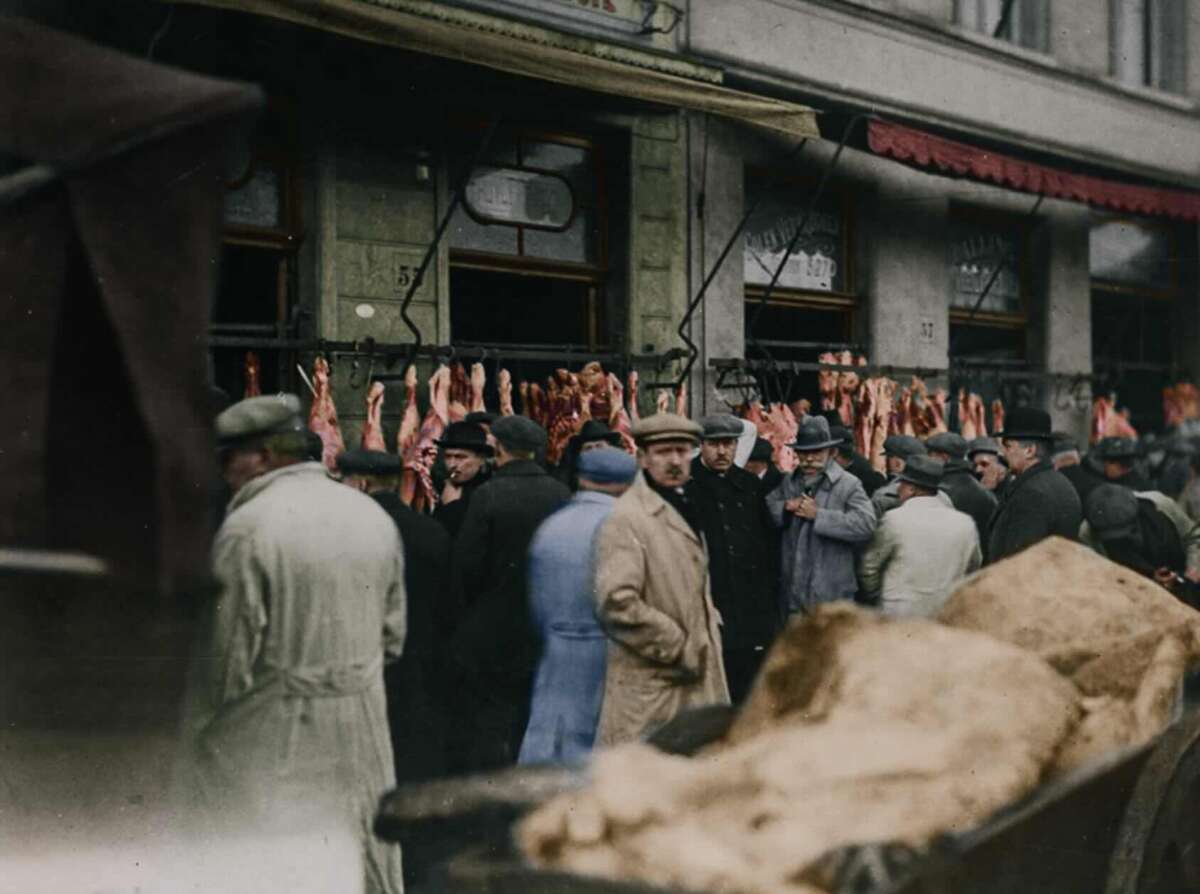 A crowd of people in coats and hats gather outside a butcher shop with meat hanging in the window along a city street. The scene appears historical, with muted colors and early 20th-century clothing.
