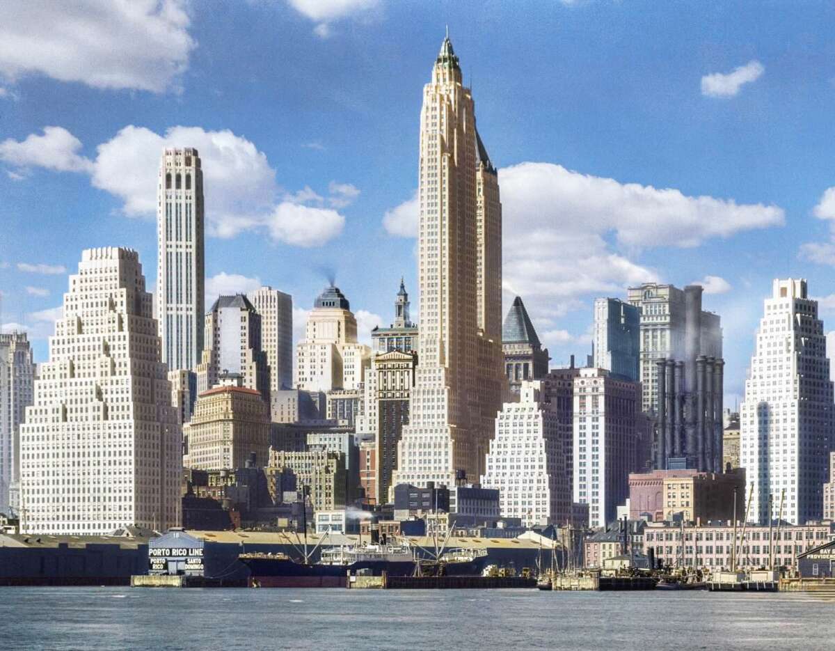 A city skyline with tall skyscrapers and historic buildings under a blue sky with scattered clouds, viewed from across a body of water.