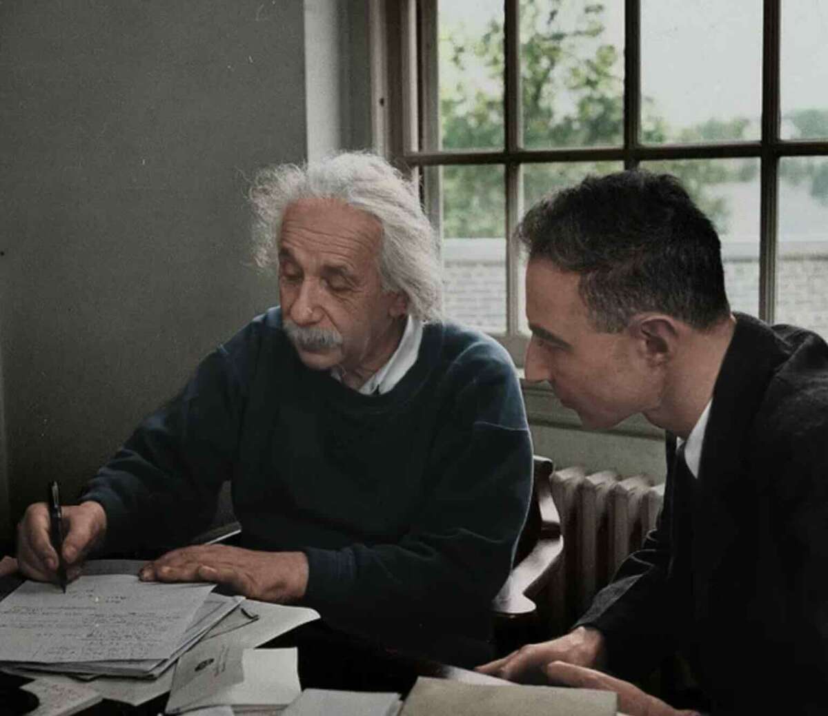 Two men sit at a desk by a window; one, with white hair and a mustache, writes on papers while the other, with dark hair, leans in closely and watches attentively.