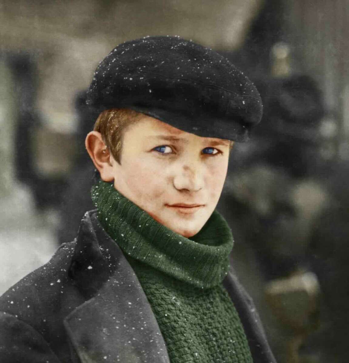 A young boy with light skin, wearing a black flat cap, green turtleneck sweater, and dark coat stands outside in falling snow, looking at the camera with a faint smile. The background is blurred.
