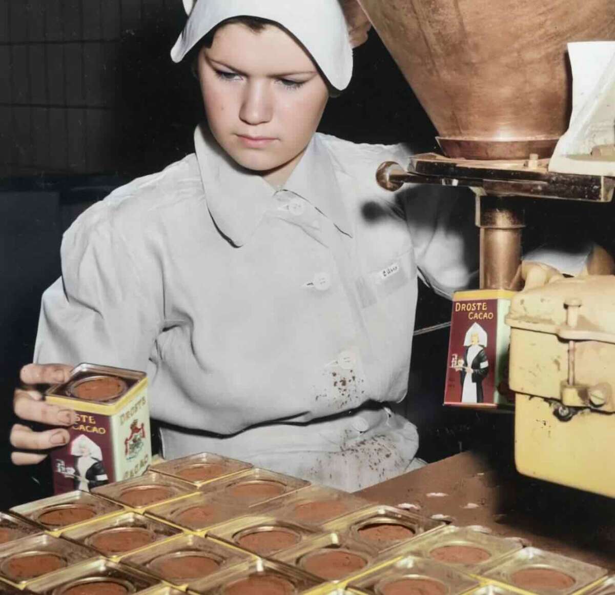 A woman in a white uniform and headscarf fills metal tins with cocoa powder at a production line. She holds a tin labeled "Droste Cacao" while other tins are arranged in rows on the work surface.