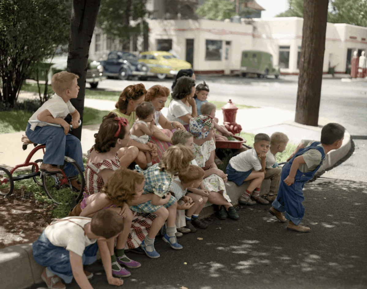 A group of young children in colorful 1950s clothing sit and crouch on a curb by a tree, watching a boy walking in front of them on a sunny day in a suburban neighborhood with cars in the background.