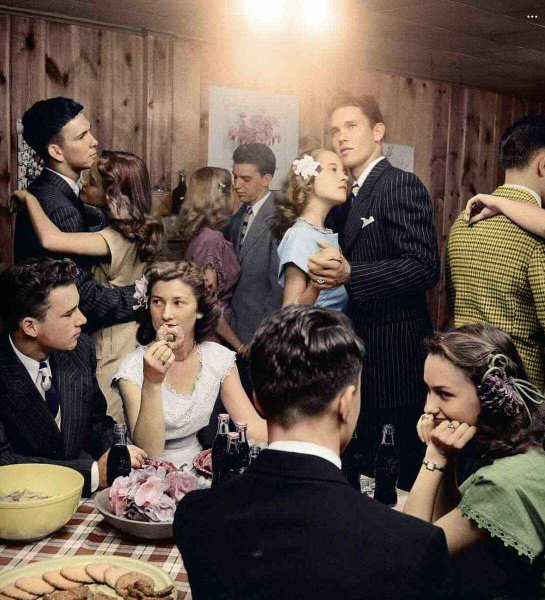 A group of young adults in vintage attire dance and sit around a table with snacks and sodas in a warmly lit, wood-paneled room, creating a lively, nostalgic atmosphere.
