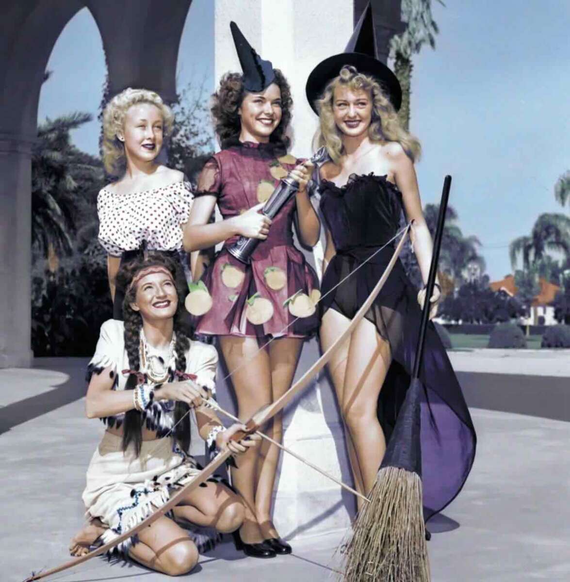 Four women in costumes pose outdoors; one is dressed as a Native American with a bow, another as a princess, and two as witches, one holding a broom and the other a wand. They are smiling and standing under an archway.