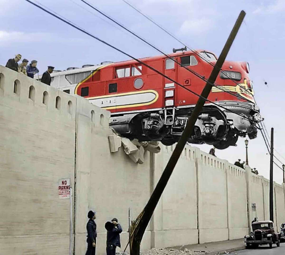 A red and silver train hangs precariously over the edge of a tall concrete wall, with people observing from above and below. A leaning utility pole and a vintage car are visible near the wall's base.