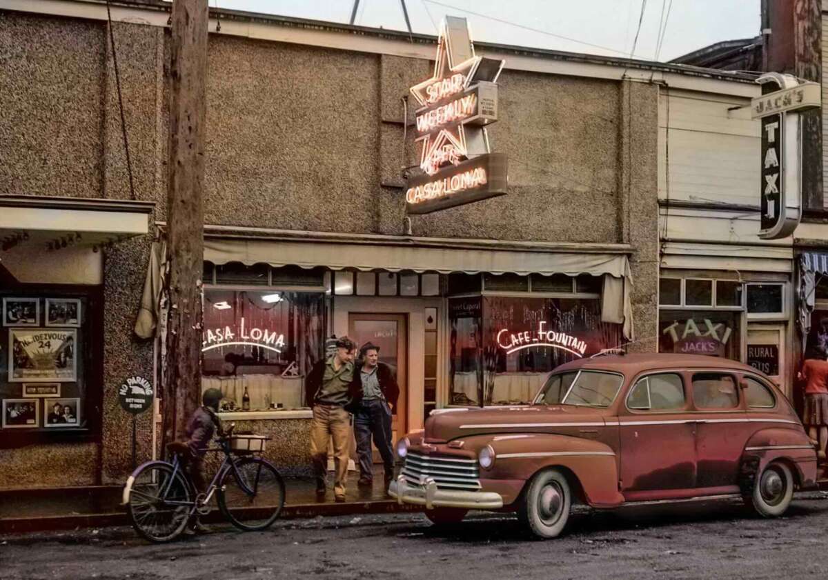 Two people stand outside a vintage café with a neon sign reading "CASA LOMA." A classic car and a bicycle are parked out front. The scene has an old-fashioned, nostalgic atmosphere, suggesting a mid-20th-century setting.