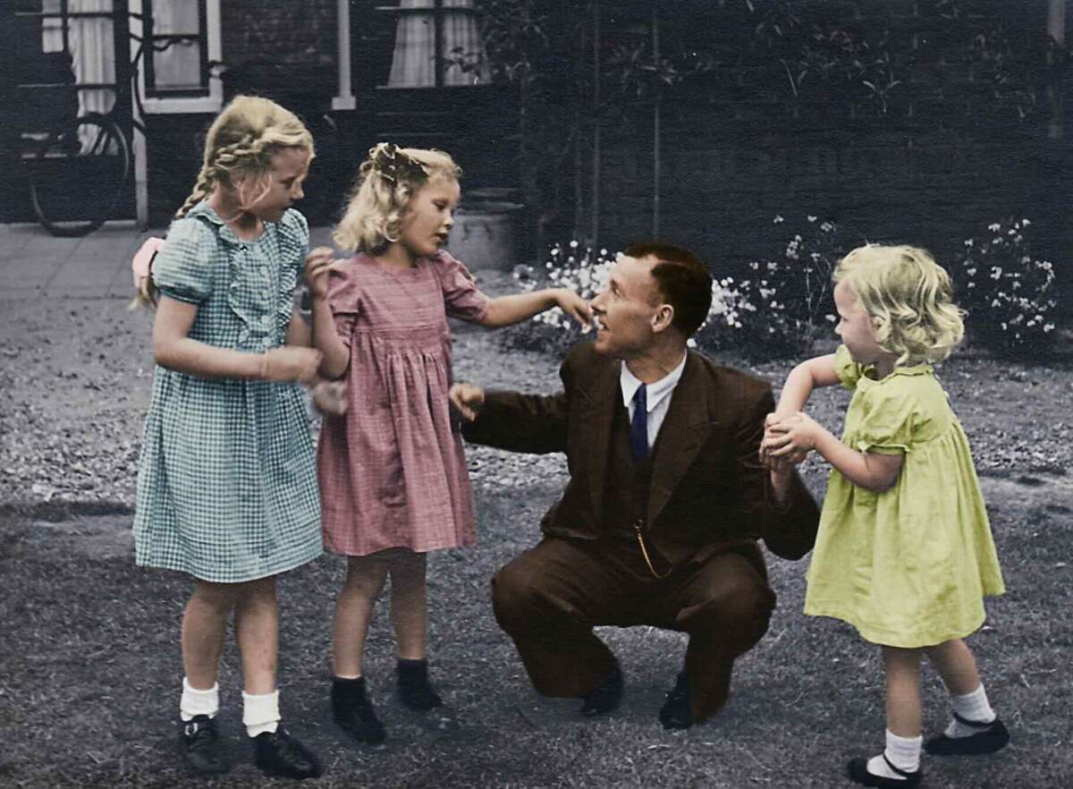 A man in a suit kneels on grass, smiling and holding hands with three young girls in colorful dresses—green, pink, and yellow—in a garden with flowers and a house in the background.