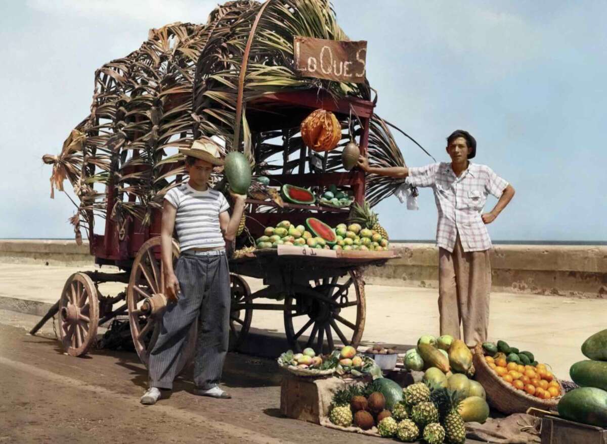 Two men stand by a colorful fruit cart decorated with palm leaves, displaying watermelons, pineapples, bananas, and other produce by a roadside under a clear sky.