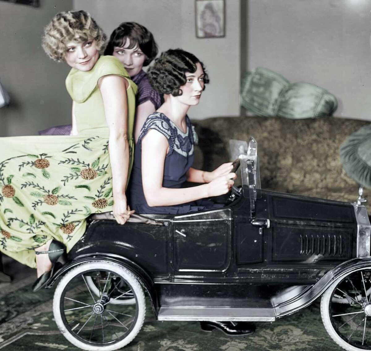 Three young women from the early 20th century pose playfully with a vintage toy car indoors; one sits inside while two others perch on the back, all smiling and wearing period dresses.