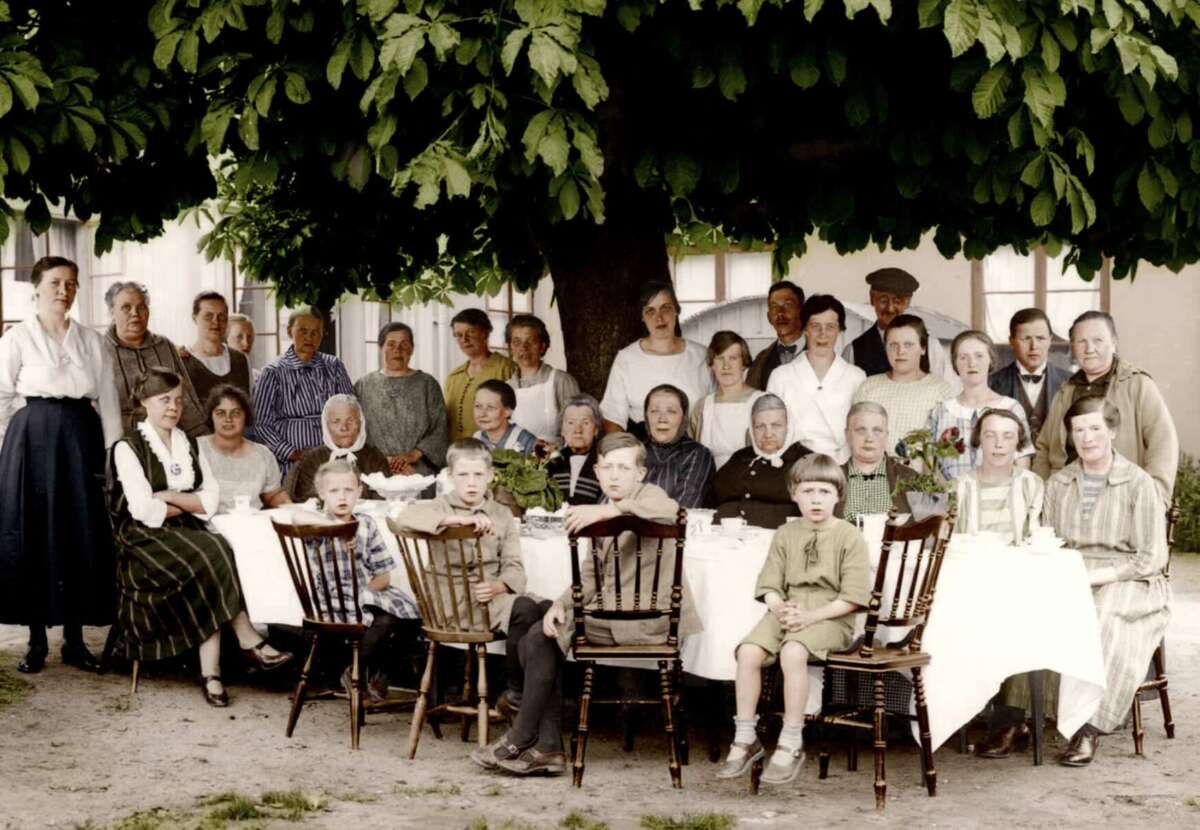 A large group of people, including men, women, and children, sit and stand around a long, outdoor table under a tree, posing for a photo. The scene looks vintage, with everyone dressed in early 20th-century clothing.
