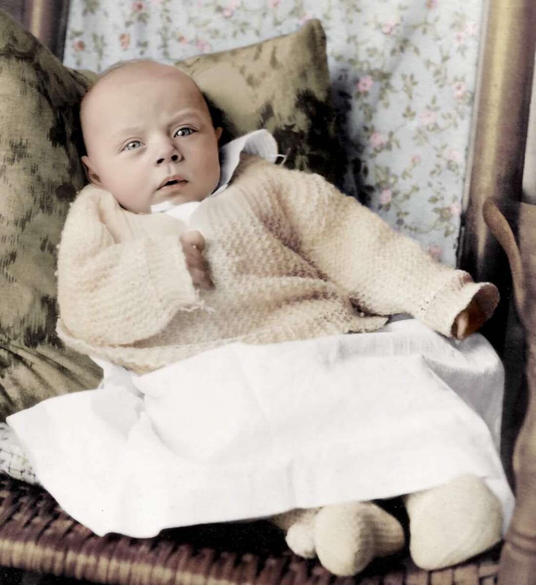 A baby wearing a light-colored knitted sweater and white blanket sits propped up on cushions with a floral wallpaper background. The baby has a serious expression and is looking slightly off camera.
