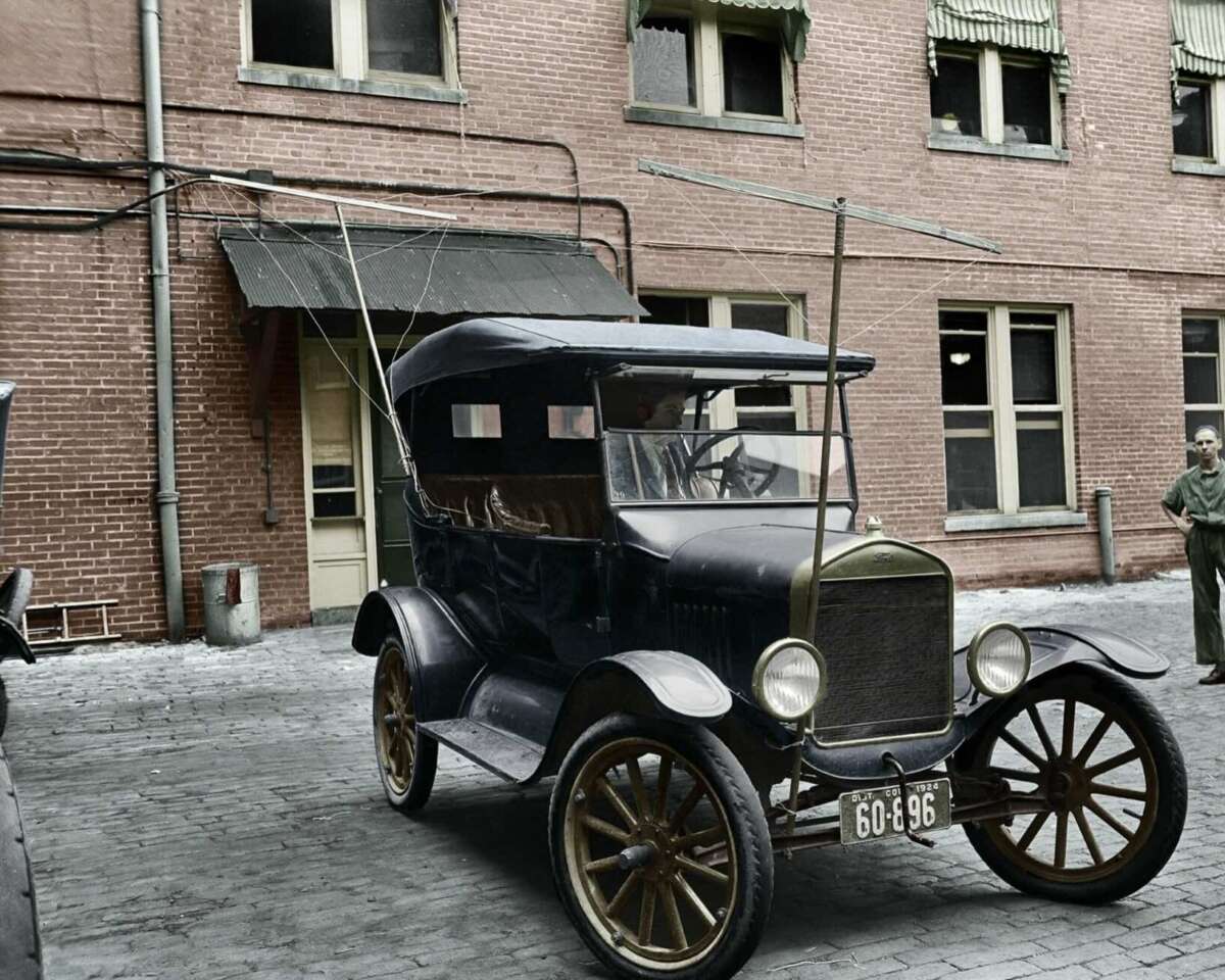 A vintage black car with a convertible roof and wooden-spoked wheels is parked on a cobblestone street in front of a brick building. A man stands nearby, and metal rods extend upright from the car.