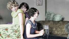 Three women in vintage clothing sit closely together indoors, with one woman appearing to drive and the others posing playfully behind her. The setting includes a couch with green pillows in the background.