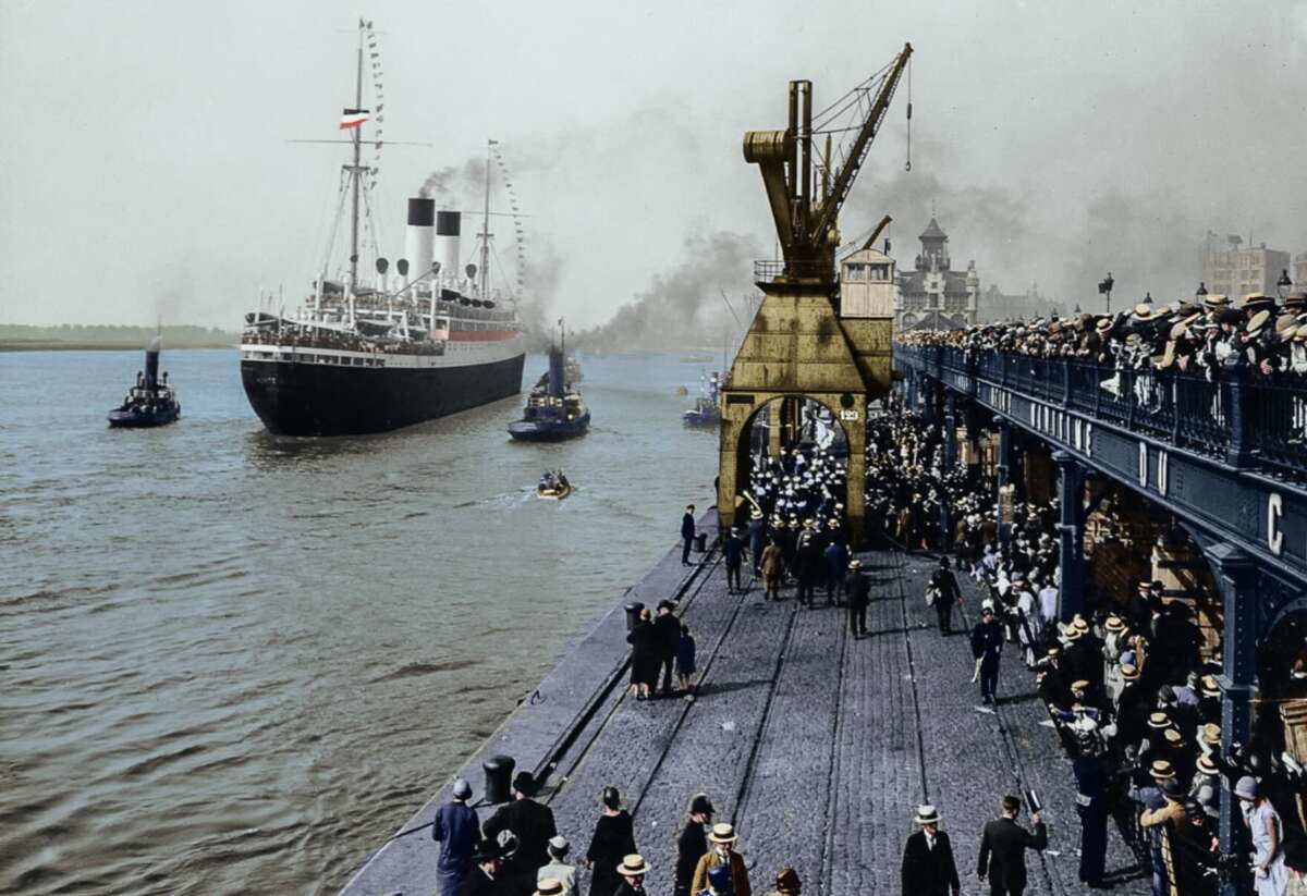 A large, early 20th-century ocean liner departs a crowded port; people in period clothing stand on the pier and balconies, watching as tugboats guide the ship down the river under a cloudy sky.