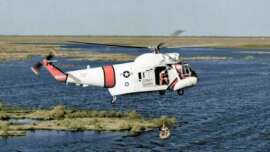 A U.S. Coast Guard helicopter hovers above water as a rescuer lowers a basket to lift a person to safety. The landscape is flat with patches of grass and water.