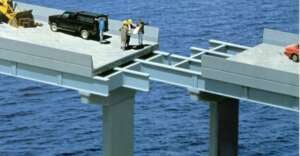 A group of people in hard hats stand on an unfinished bridge over water, with a black SUV behind them. The bridge segments do not align, leaving a large gap between them. A red car is on the opposite section.