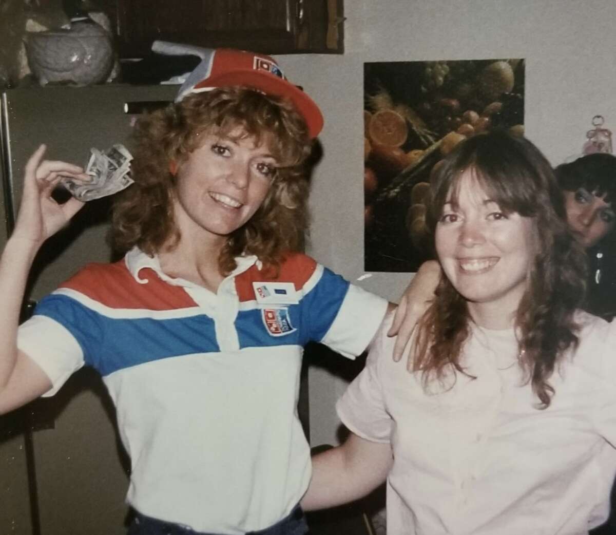 Two smiling women pose indoors; one wears a red, white, and blue Domino’s Pizza uniform and hat, holding cash in one hand, while the other wears a light pink shirt. A third person peeks from behind them. A fruit poster is on the wall.