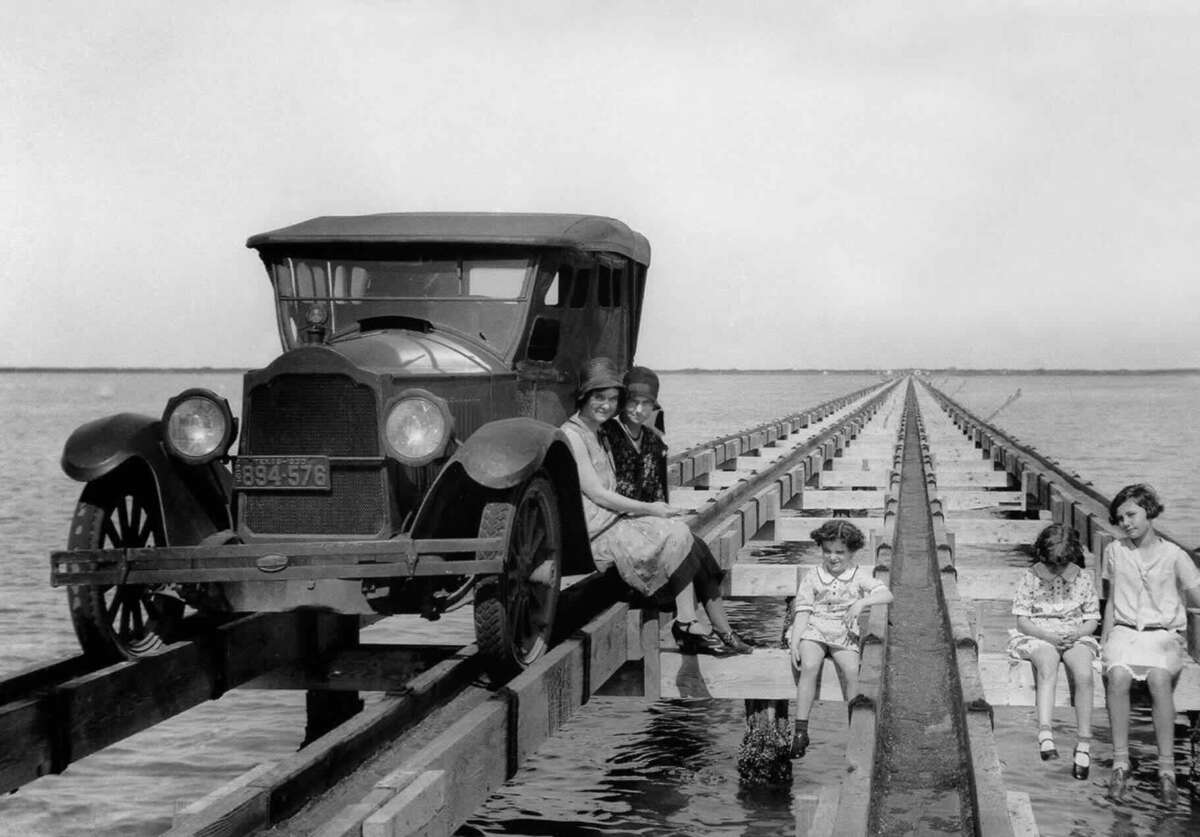 A vintage car and several women sit on narrow wooden tracks over water; three women perch beside the car, while three young girls dangle their feet above the water further down the tracks.
