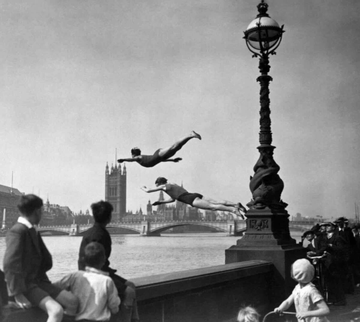 Two boys in swimsuits dive off a bridge into the River Thames, as people watch. The Houses of Parliament and Big Ben are visible in the background on a sunny day.