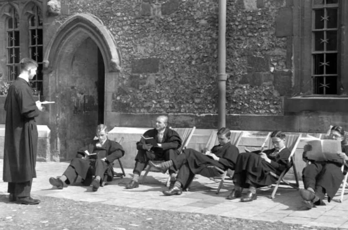 A group of students in black academic gowns sit on deck chairs outside an old stone building, reading or writing, while a standing teacher in similar attire speaks to them.