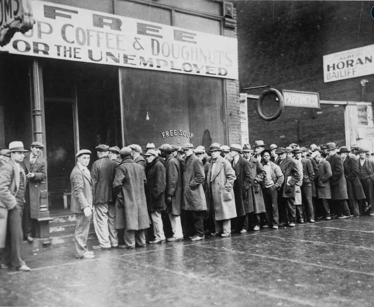 A long line of men in coats and hats waits outside a building with a sign offering "Free Soup, Coffee & Doughnuts for the Unemployed" during the Great Depression era. The street and surrounding area look bleak and crowded.