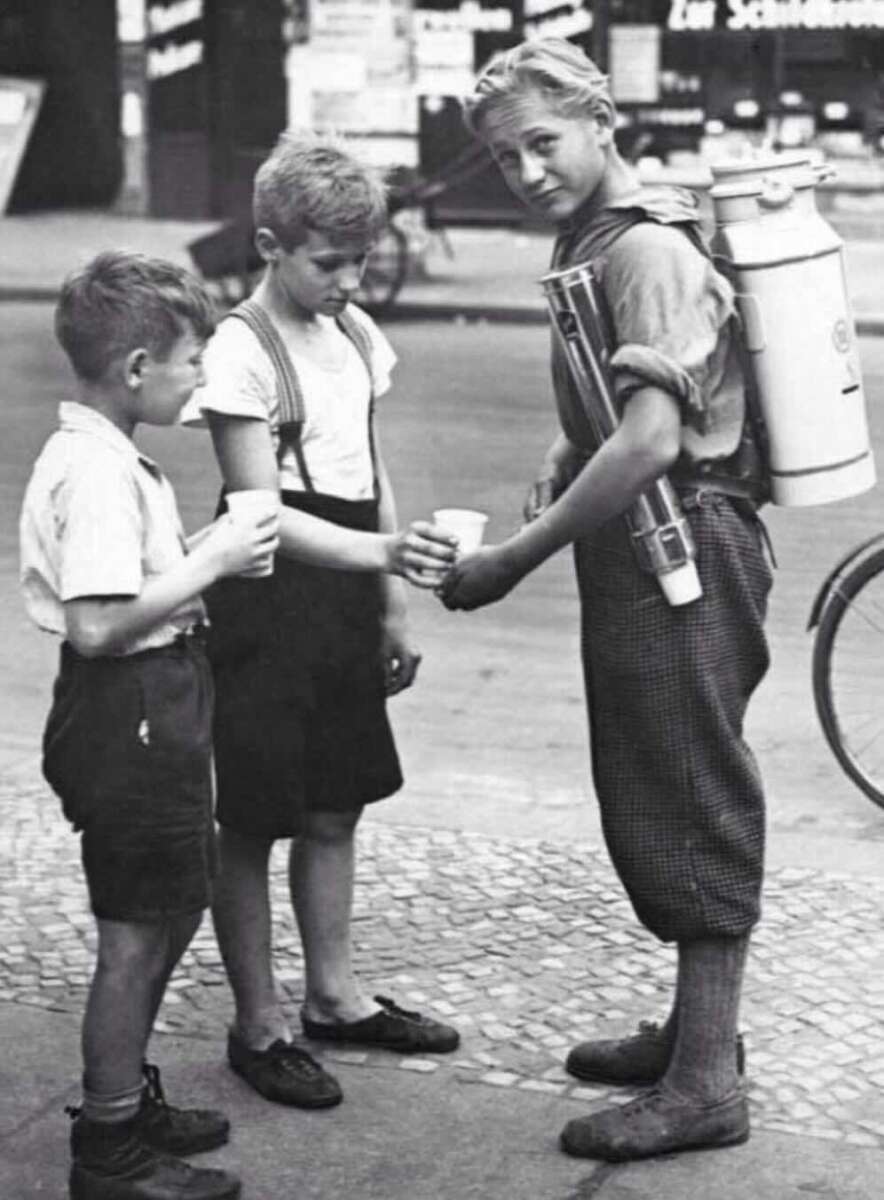 A boy carrying a milk can and schoolbooks pours milk into a cup held by one of two younger boys on a city sidewalk, all dressed in vintage clothing.