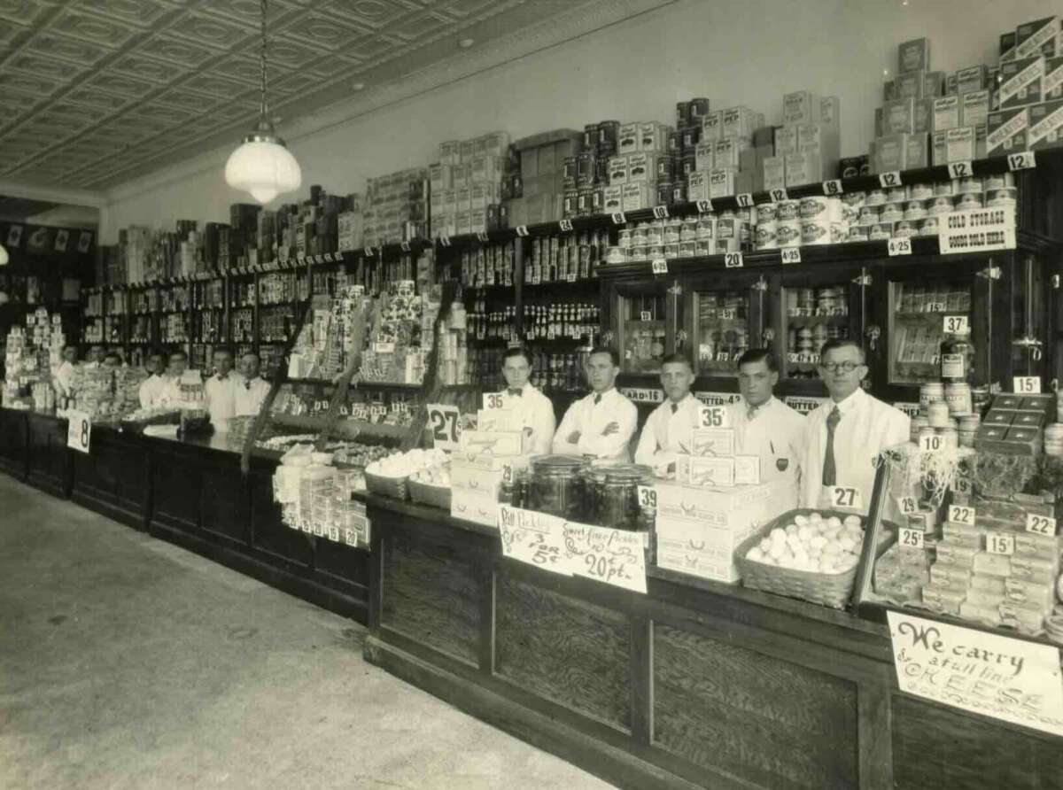 A black-and-white photo of a grocery store with six men in white coats standing behind a counter, surrounded by shelves stacked high with canned and boxed goods and displays of produce and eggs. Handwritten signs show prices and specials.