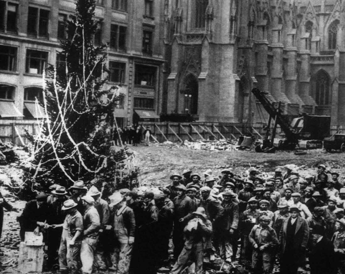 A large group of construction workers gather around a decorated Christmas tree in a city construction site with tall buildings and a church in the background; a crane is visible nearby. The scene is in black and white.