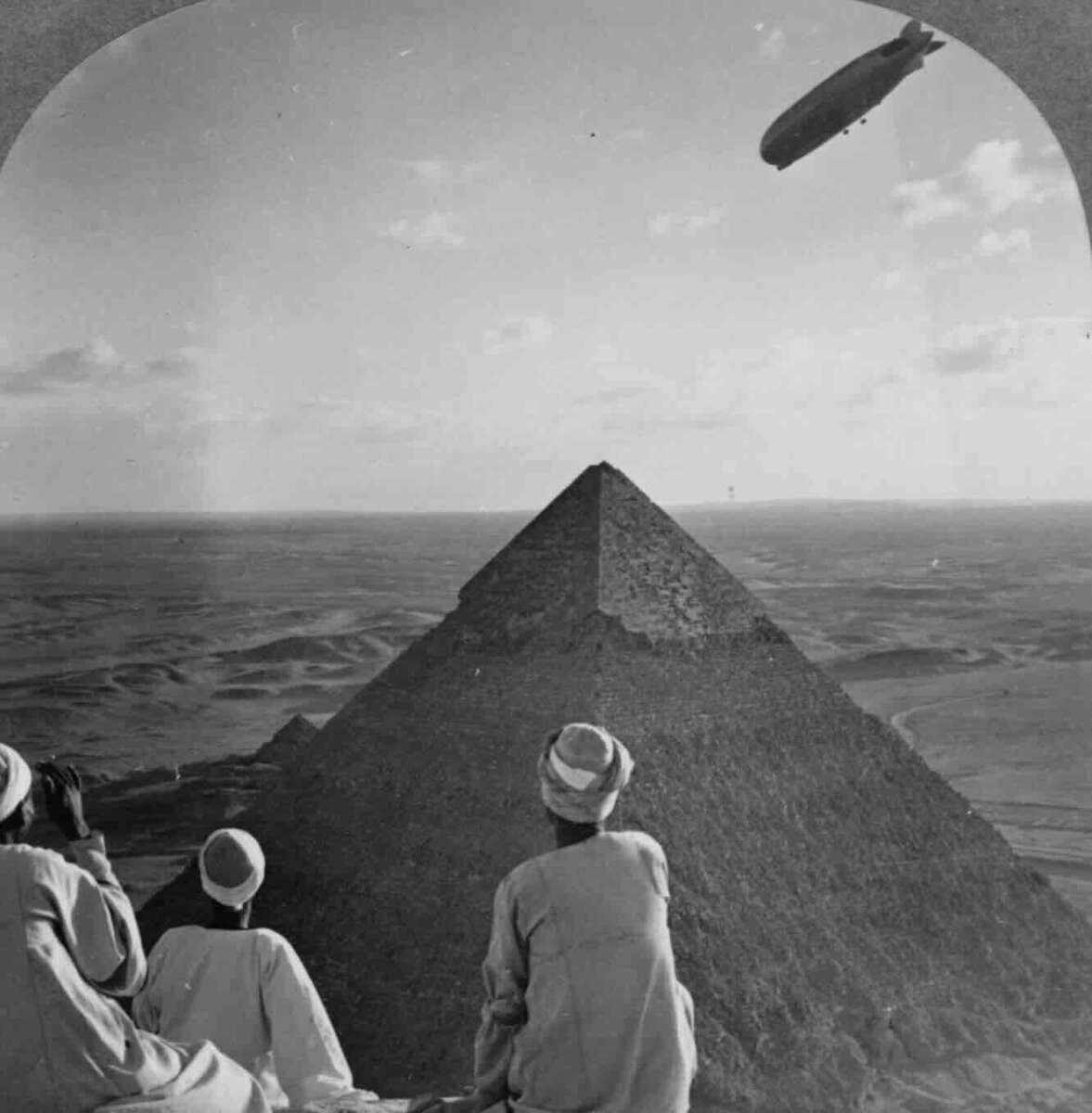 Three people in traditional clothing sit atop a pyramid, looking out over the desert as a large airship floats in the sky above another pyramid in the distance.