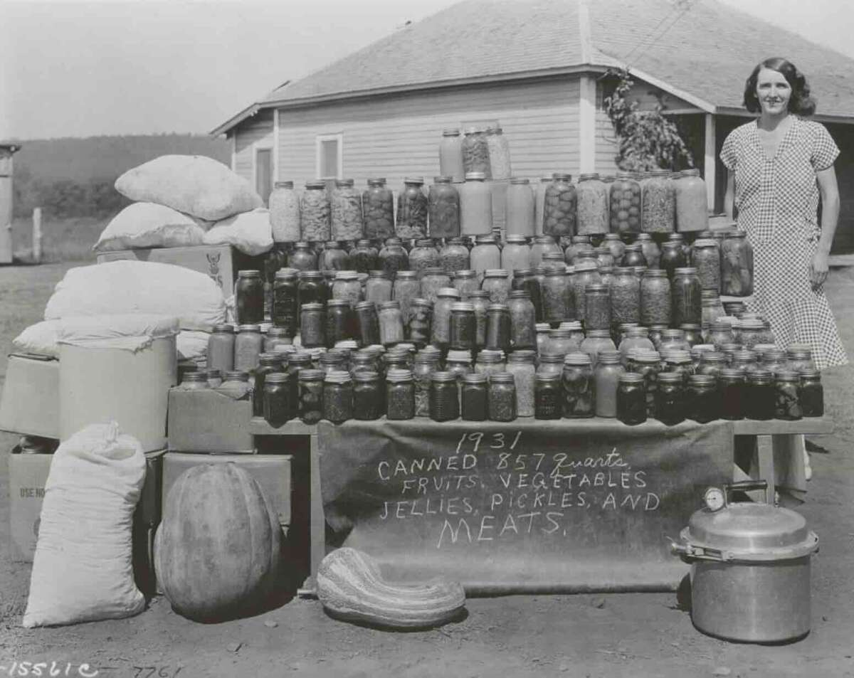 A woman stands beside a display of jars filled with preserved fruits, vegetables, jellies, pickles, and meats, labeled “1931 Canned 857 Quarts,” with sacks, a large squash, and a house in the background.