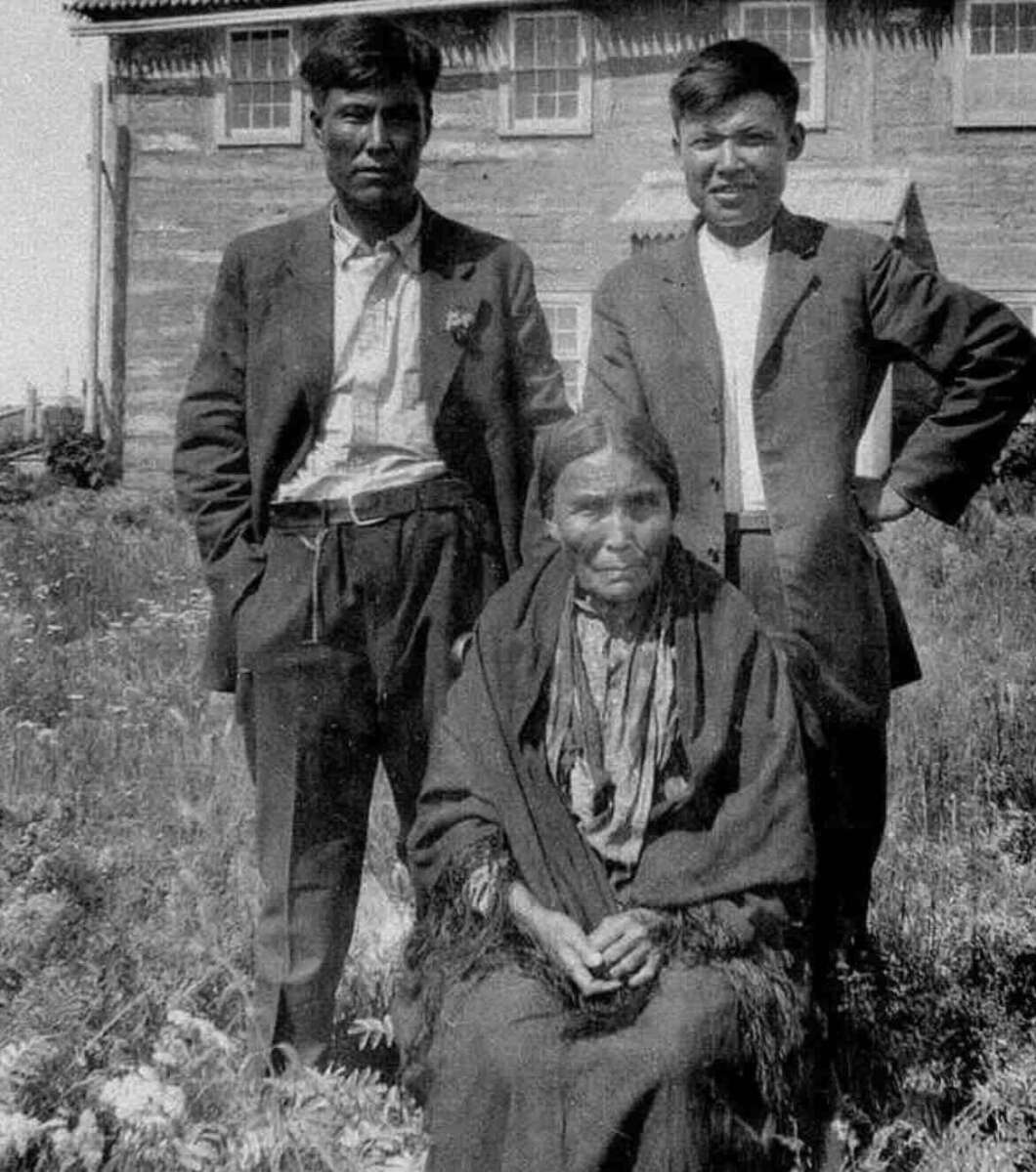Three people pose outdoors in front of a wooden building: two men standing in suits and a woman seated in front wearing a shawl. Grass and wildflowers grow around them, and the scene appears old-fashioned or historical.