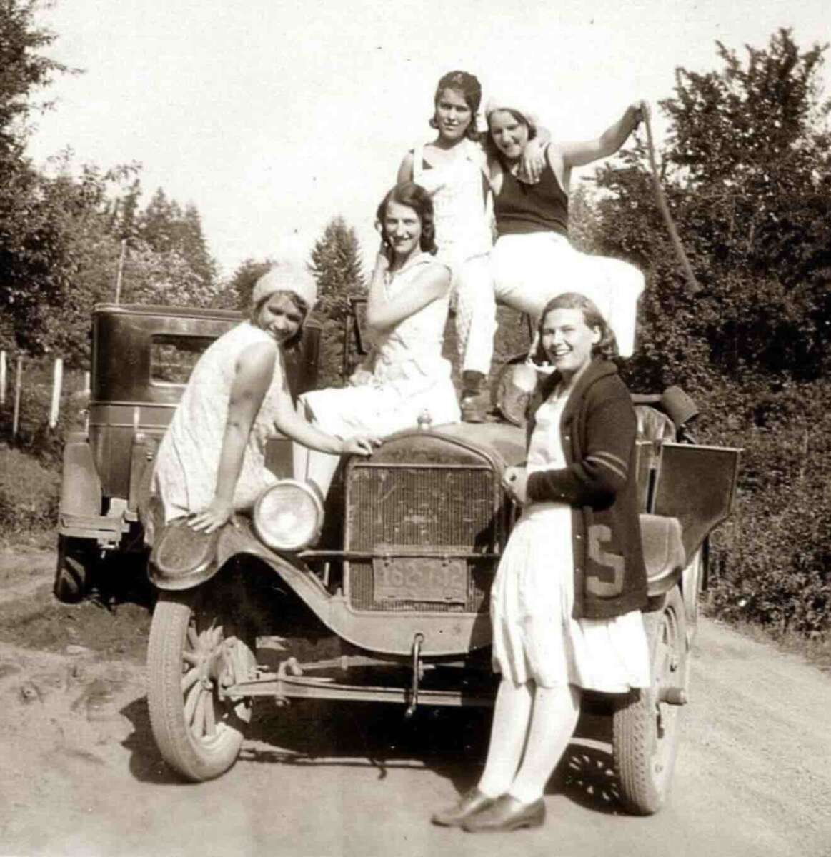 Five young people in 1920s clothing pose and smile on and around an old car parked on a dirt road, surrounded by trees and another vintage car in the background.