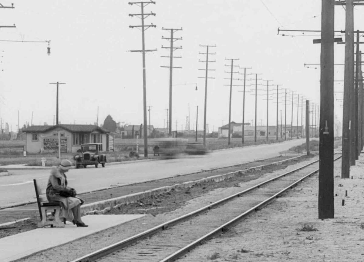 A woman sits alone on a bench beside railroad tracks, with power lines and telephone poles lining a road. An old car and a blurred vehicle are visible in the background, suggesting motion. The scene appears desolate and quiet.