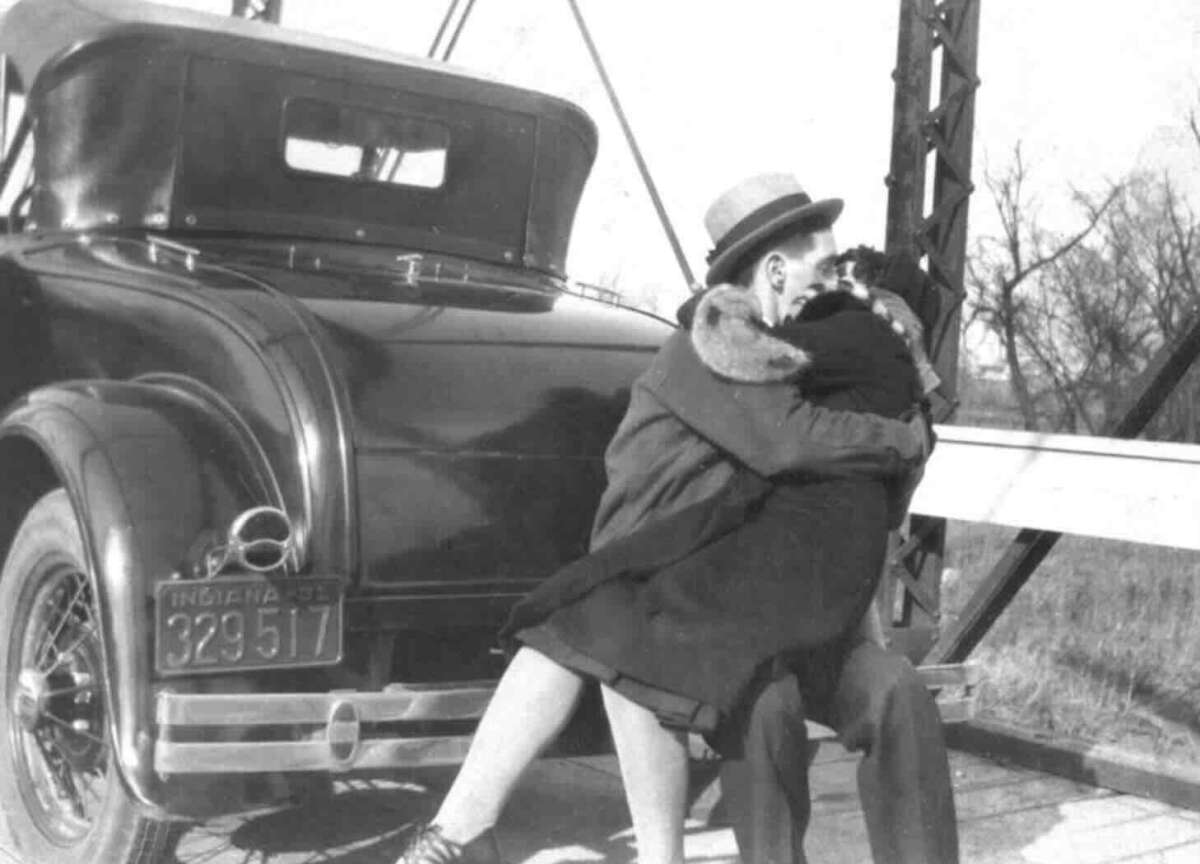 A couple sits on the bumper of an old car, embracing and kissing. The car has an Indiana license plate and is parked on a bridge. The scene is in black and white, evoking a vintage feel.