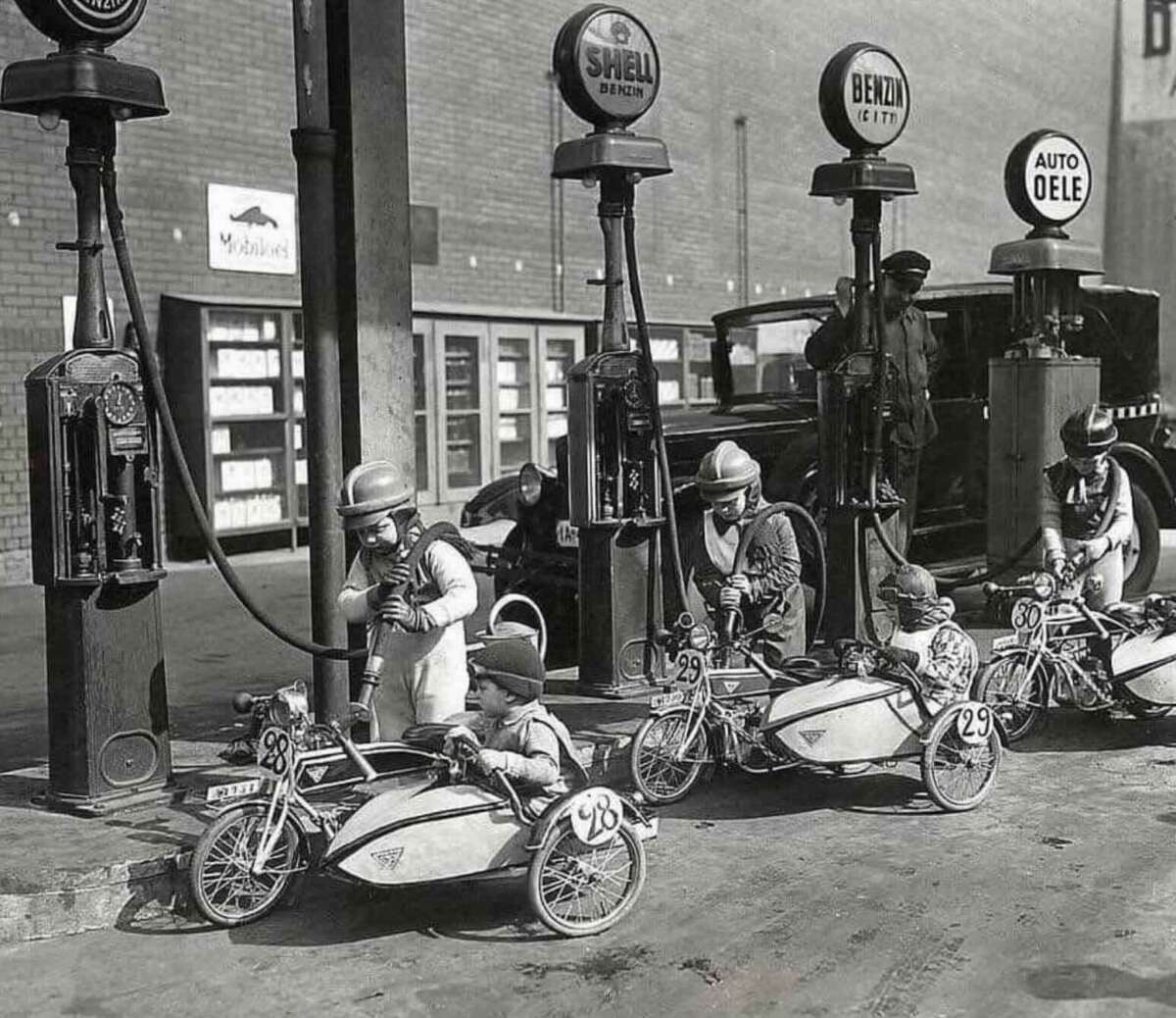 Children in vintage racing outfits sit in small pedal cars, lined up beside old-fashioned gas pumps at a service station. An adult stands nearby, and a vintage car is parked in the background. The scene appears to be from the early 20th century.