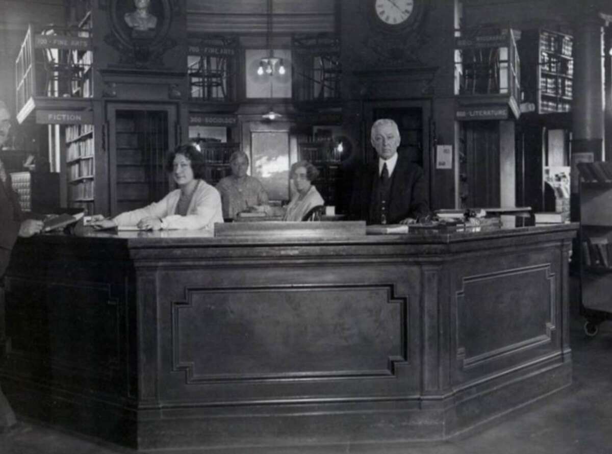 Four people sit and stand behind a large, dark wooden library desk in a vintage library. Bookshelves, lamps, and a clock are visible in the background, giving the scene an old-fashioned, scholarly atmosphere.
