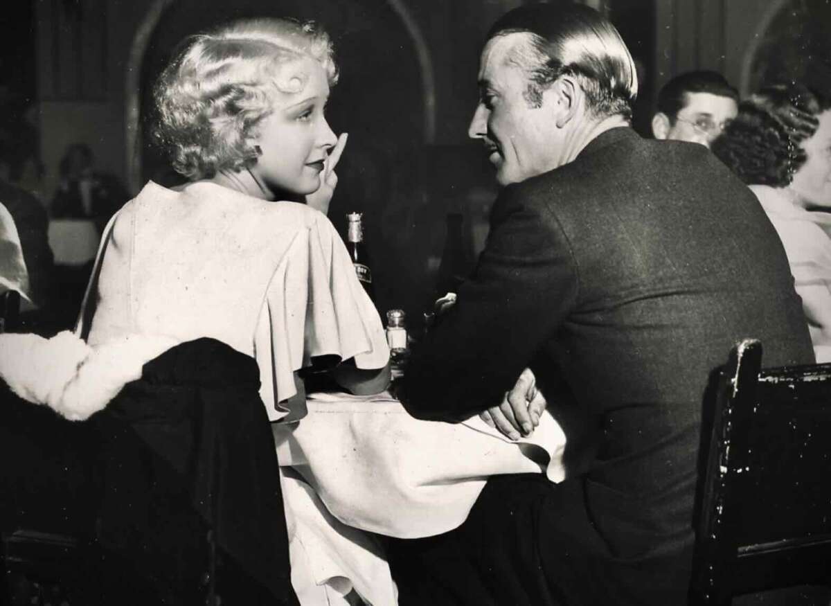 A woman with short, wavy blonde hair and a man in a suit sit closely at a table in a dimly lit restaurant, engaged in conversation, with other diners visible in the background.