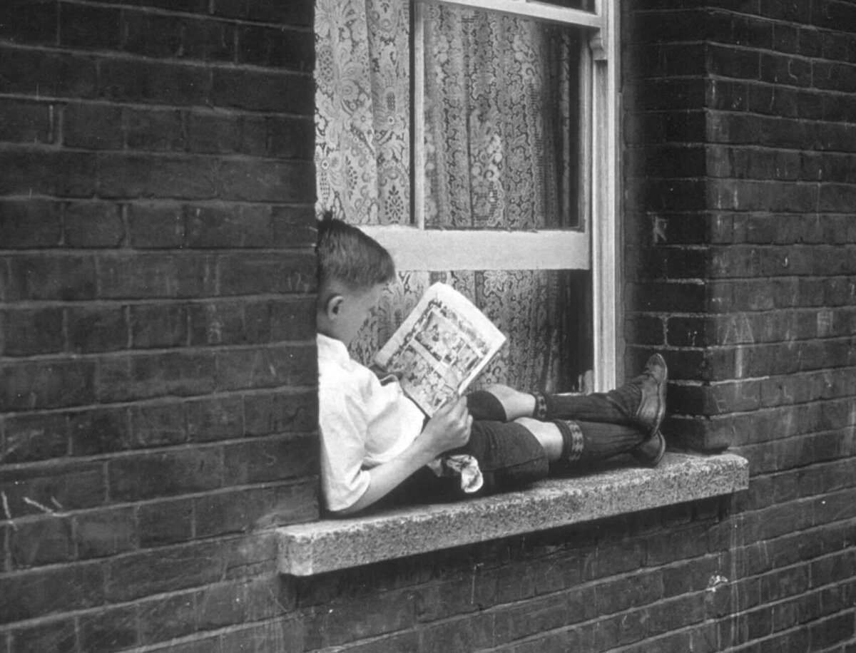 A young boy sits on a windowsill with legs stretched out, reading a comic book. The window behind him is open, and lace curtains are visible inside. The building exterior is made of dark bricks.
