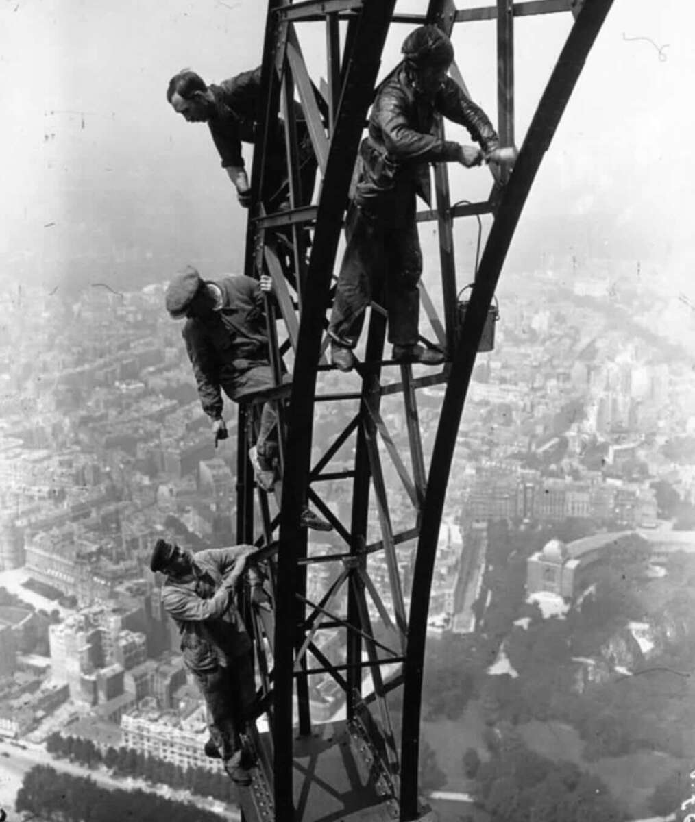 Four workers climb and repair the metal frame of a tall structure high above a city, with buildings and trees visible far below in the background.