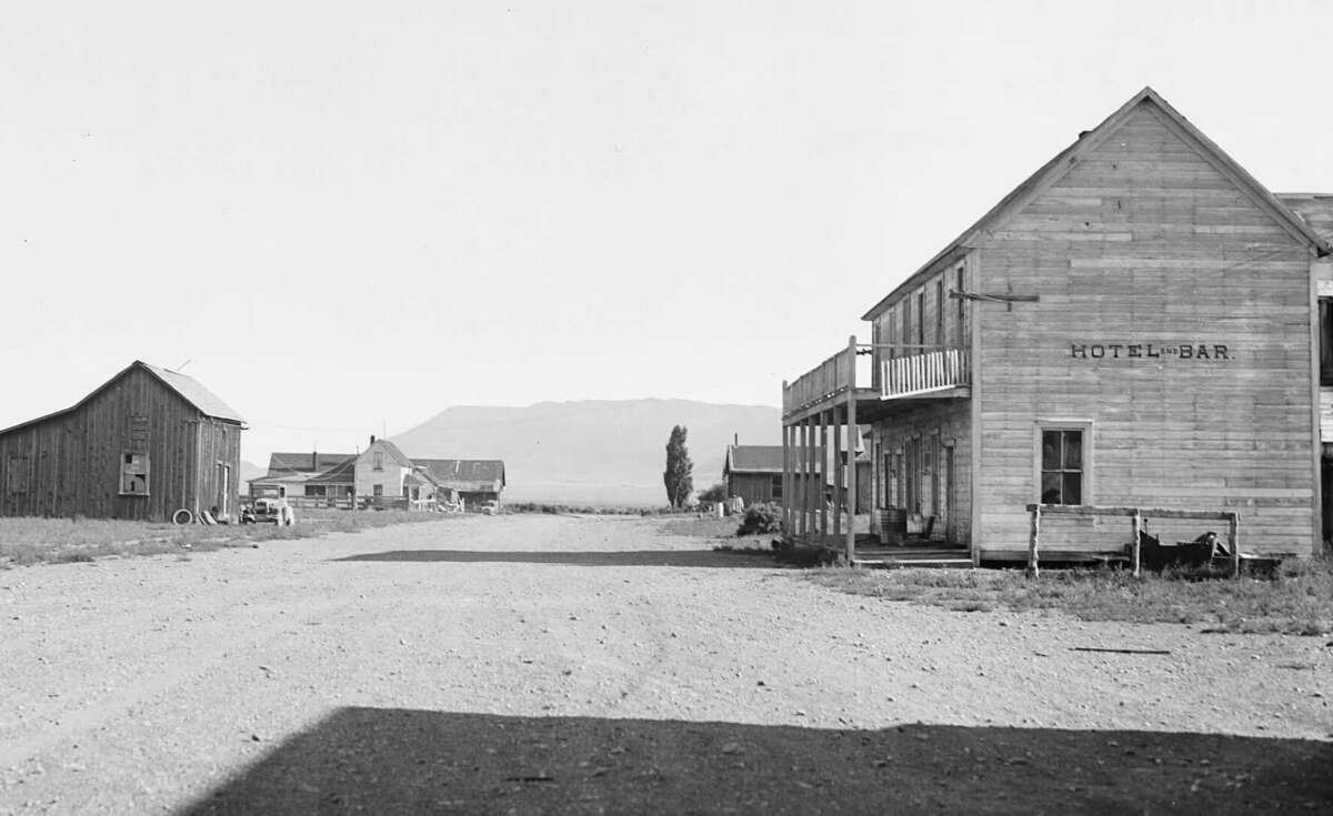 A dirt road runs through a small, deserted town with wooden buildings, including a two-story "Hotel & Bar" on the right. The scene is quiet, with distant mountains in the background.