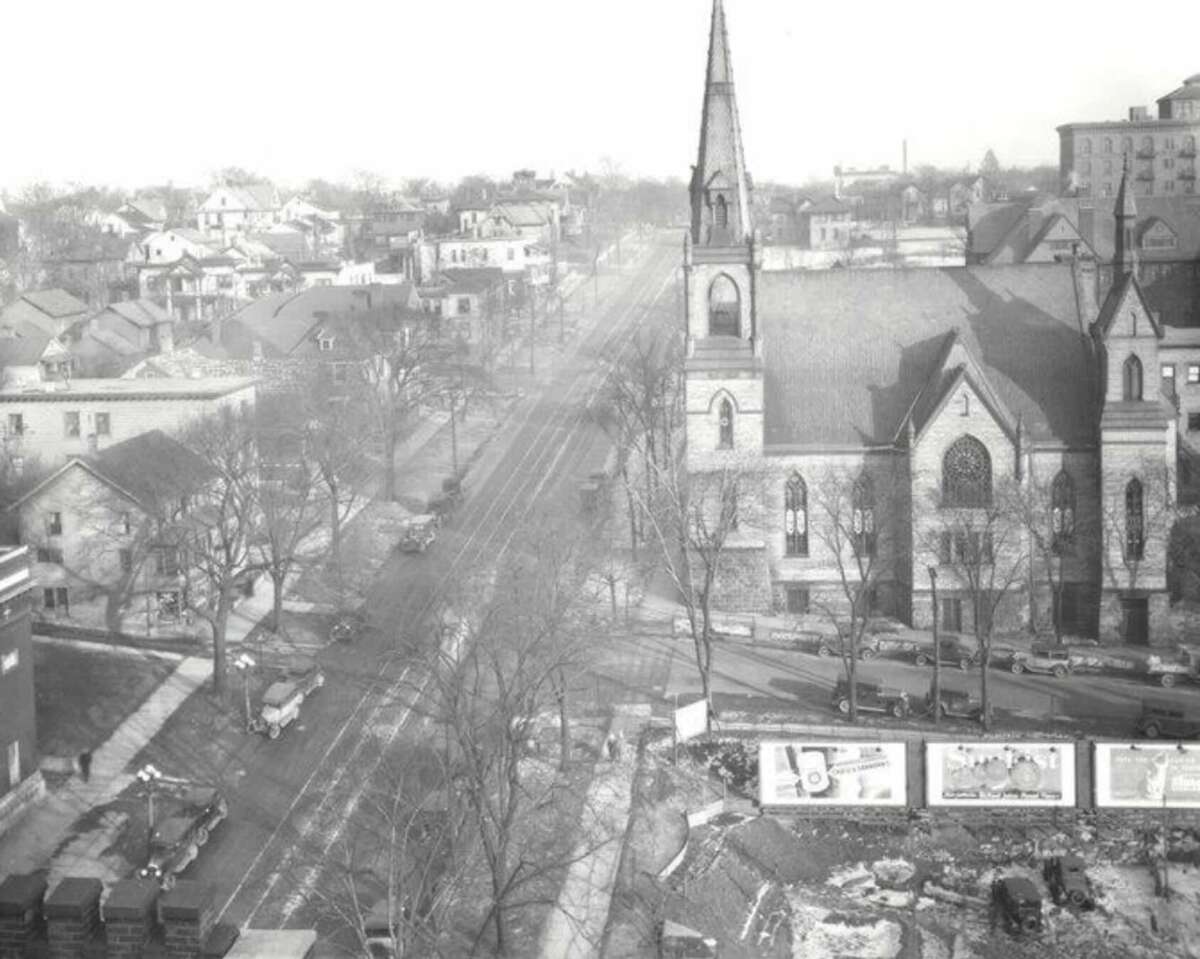 A black-and-white photo shows a city intersection with a large church featuring a tall steeple. Cars and trees line the streets, and billboards are visible in the foreground. Houses and buildings fill the background.