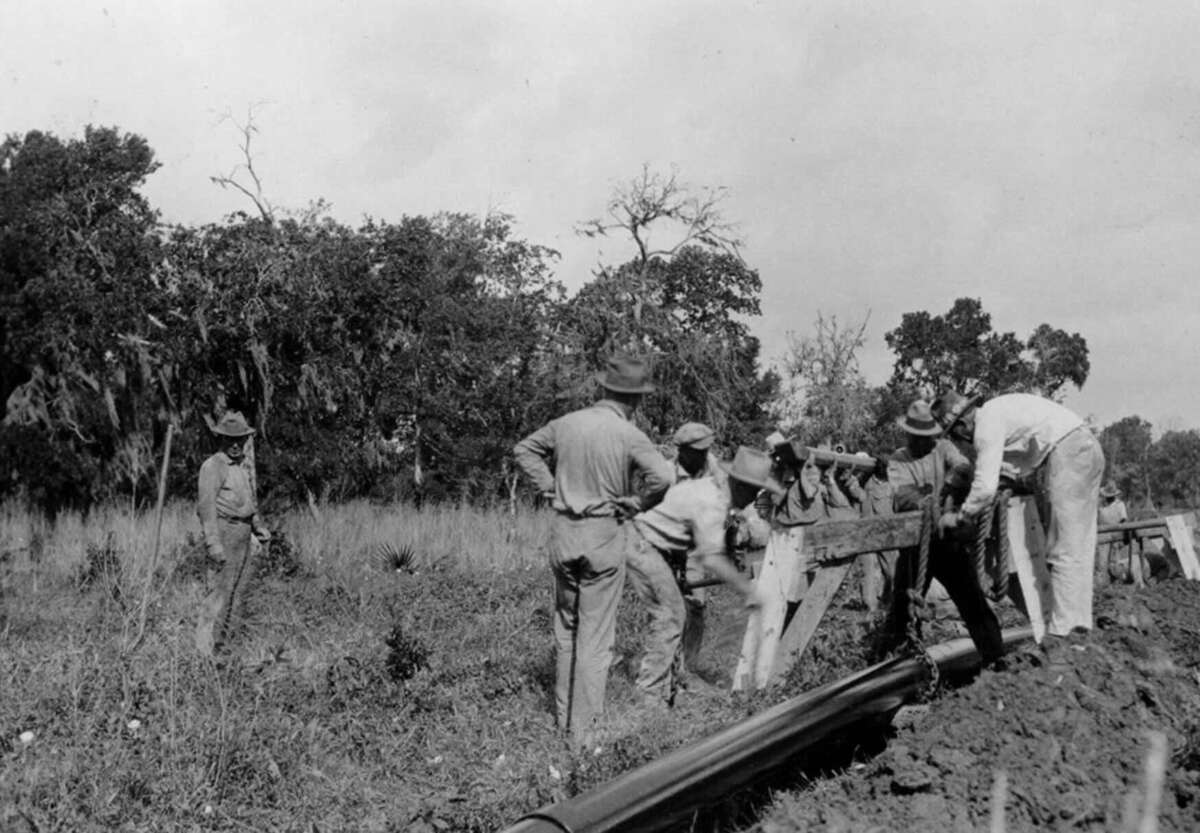 A group of workers install or repair a pipeline in a rural, grassy area with trees in the background. Several men are handling the pipe while others stand nearby, watching or assisting.
