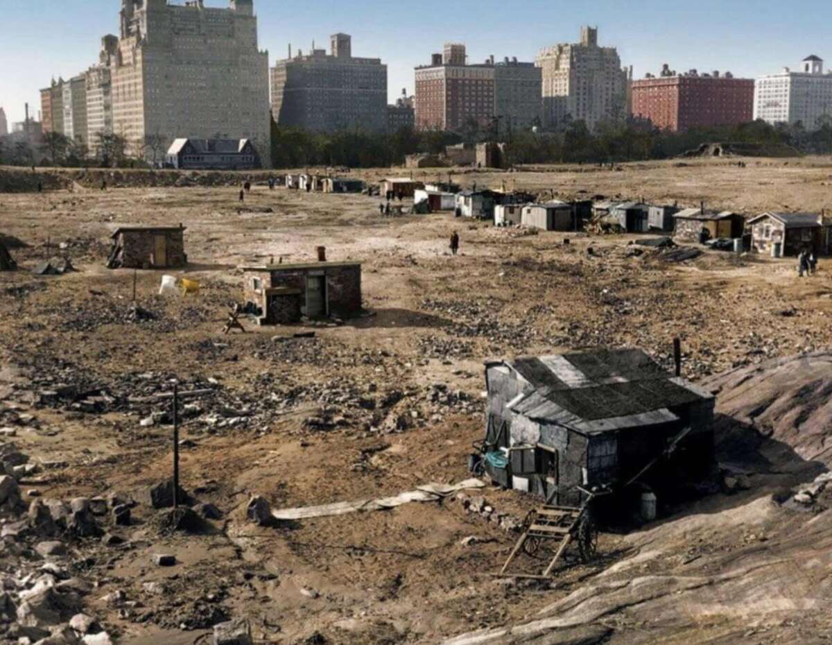 Shacks and makeshift homes stand on a barren, rocky landscape in the foreground, with a row of tall, upscale apartment buildings visible in the background under a clear sky.